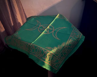 May include: A square green fabric cloth featuring a gold-colored print of a triple moon symbol with a central pentacle. The border is decorated with an intricate Celtic knotwork pattern that runs along all four edges of the textile material.