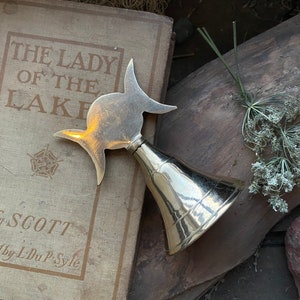 May include: A gold-colored metal bell with a triple moon design. The bell is sitting on top of a book titled "The Lady of the Lake" by Scott.