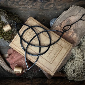 May include: A black metal Celtic knot design with two gold bells hanging from it. The knot is resting on top of a vintage book titled "The Lad of the Lake" by Scott. The book is surrounded by moss, wood, and a piece of antler.