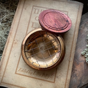 May include: A brass incense burner with a wire mesh insert and a wooden lid. The burner is sitting on an old book with the text "Edited by L.L. Pyle".