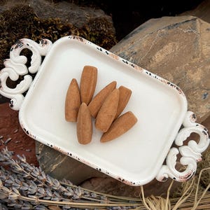 May include: A white rectangular tray with ornate handles holds several brown incense cones. The tray has a distressed, vintage look with dark accents. Dried lavender and other natural elements surround the tray, suggesting a focus on aromatherapy or home fragrance.