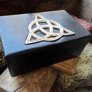 May include: A black wooden box with a silver Celtic knot design on the lid. The box is sitting on a wooden surface.