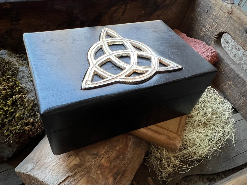 May include: A black wooden box with a silver Celtic knot design on the lid. The box is sitting on a wooden surface.