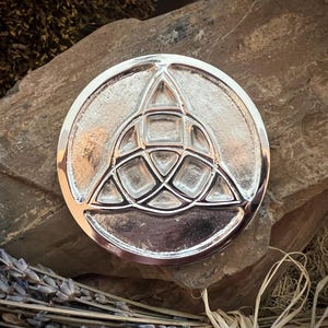 May include: A silver-colored, circular pendant with a raised Celtic knot design. The pendant is set against a natural stone background, with dried lavender and other natural elements in the foreground.