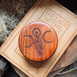 May include: A wooden round box with a triple moon and a spiral design carved into the top. The box is made of dark wood with a natural finish.