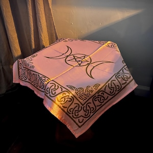 May include: A square light pink fabric cloth featuring a dark brown printed design. The center displays a triple moon symbol with a central pentagram. The border is decorated with an intricate, repeating Celtic knotwork pattern on all four sides.