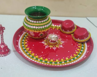 May include: A red metal plate with a green and gold rim, decorated with white pearls and gold dots. A green and gold pot with a green lid sits on the plate, along with two smaller red bowls and a silver diya.