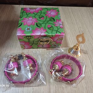May include: A set of pink silk thread bangles and earrings with gold accents, packaged in a green floral box with a silver bell.