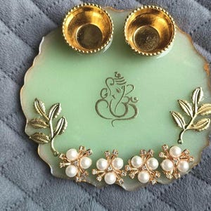 May include: A round, light green resin tray with a gold-colored Ganesha symbol in the center. The tray features two small gold-colored circular bowls at the top, two gold-colored leaf accents on the sides, and four floral pearl embellishments.