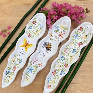 May include: Three white ceramic incense holders with a wavy design. Each holder features a different floral pattern with butterflies and bees. The holders are decorated with green, blue, red, yellow, and orange colors.