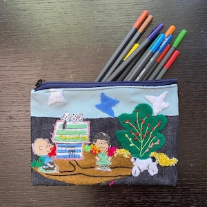 May include: A blue denim pencil case with a zipper closure. The case features an embroidered design of the Peanuts characters, including Snoopy, Woodstock, and Charlie Brown. The case is filled with colored pencils.