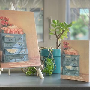 以下が含まれることがあります： イーゼルにかけられた2枚の絵画。どちらも青い引き出し付きのドレッサーを描いています。上の絵画はドレッサーの上に花が飾られており、下の絵画はドレッサーの上に赤と白のストライプの毛布が置かれています。