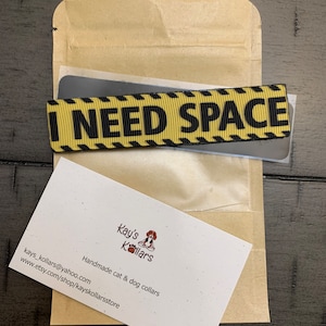 May include: A yellow and black ribbon with the text "I NEED SPACE" printed on it. The ribbon is packaged in a brown paper envelope with a business card that reads "Kay's Kollars Handmade cat & dog collars" with a website address and social media icons.