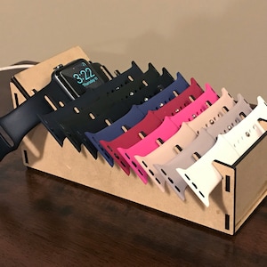 Puede incluir: Un organizador de madera para correas de Apple Watch que muestra un Apple Watch negro y varias correas de colores. Las correas son de varios colores, incluyendo negro, azul, rosa y blanco. El reloj muestra la hora 3:22.