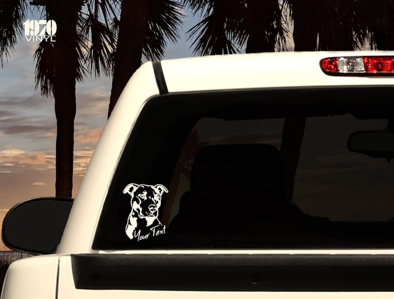 May include: White vinyl decal of a pit bull dog's head with the text "Your Text" underneath. The decal is on the back window of a white pickup truck.