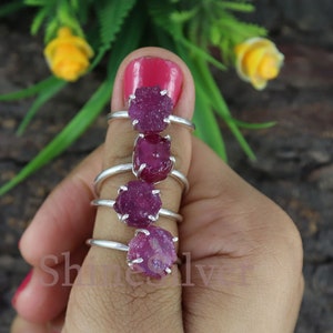 以下が含まれることがあります： 4つのシルバーリング。それぞれに未加工のルビーの宝石がセットされています。ルビーは鮮やかなピンクがかった赤色です。リングは手の上に展示されており、宝石が際立っています。背景は天然木の表面です。