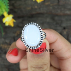 May include: A white oval gemstone ring with a silver band and a beaded border. The ring is on a finger.
