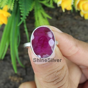 May include: A silver ring featuring a large, oval-shaped, faceted ruby gemstone. The ruby is a deep red colour. The ring is on a finger.