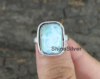 Könnte beinhalten: Ein silberner Ring mit einem rechteckigen, hellblauen Edelstein. Der Edelstein hat ein wolkiges, marmoriertes Muster. Der Ring ist in einer silbernen Fassung mit einem dekorativen Rand gefasst.