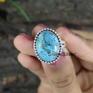 Peut inclure: Une bague en argent avec une grande pierre ovale en turquoise sertie dans une b&eacute;li&egrave;re &agrave; perles. La pierre est de couleur bleu et vert avec des paillettes dor&eacute;es.