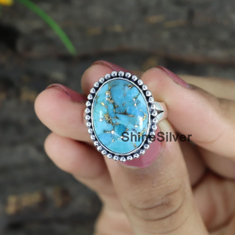 Peut inclure: Une bague en argent avec une grande pierre ovale en turquoise sertie dans une b&eacute;li&egrave;re &agrave; perles. La pierre est de couleur bleu et vert avec des paillettes dor&eacute;es.
