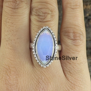 May include: A silver ring featuring a large, oval-shaped, pale blue gemstone. The gemstone is surrounded by a twisted rope-like border. The ring is on a finger. The text "ShineSilver" is visible on the ring.