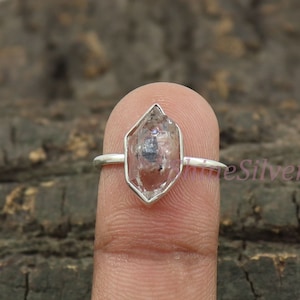 Puede incluir: Un anillo de plata con un cristal de cuarzo transparente en forma de diamante. El cristal está engastado en un bisel de plata y el anillo está en un dedo.