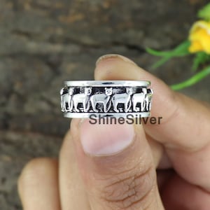 May include: A silver ring featuring a design of a row of cats walking around the band. The ring is on a finger.