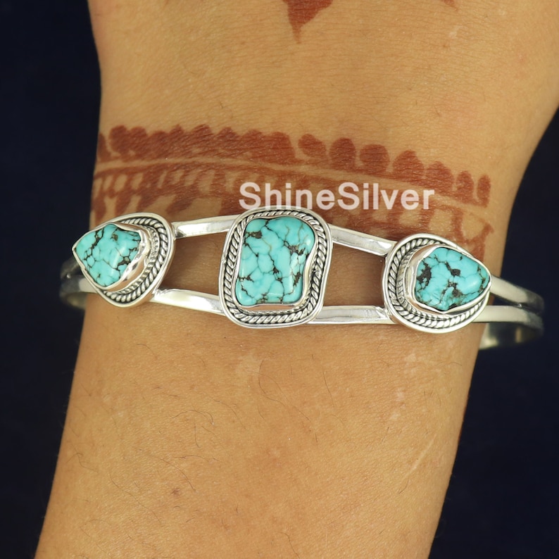 K&ouml;nnte beinhalten: Silbernes Armreif-Armband mit drei T&uuml;rkis-Steinen. Die Steine sind in silbernen Fassungen gefasst und von einem silbernen Seil-Design umgeben. Das Armband ist offen und hat ein Scharnier.