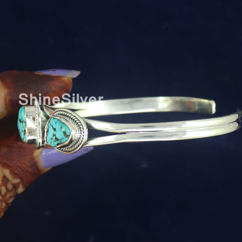 K&ouml;nnte beinhalten: Silberner Armreif mit zwei t&uuml;rkisfarbenen Steinen, die in Silber gefasst sind. Das Armband ist offen und hat ein schlichtes Design.