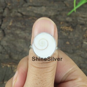 May include: A silver ring with a white shell inlay featuring a spiral pattern. The ring is on a person's finger.
