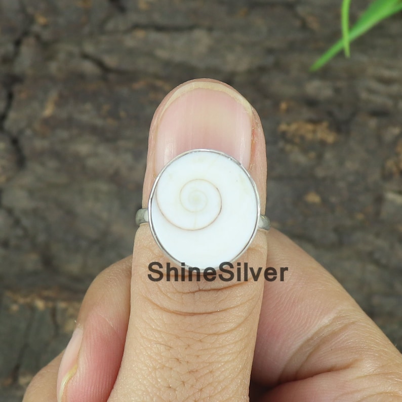 May include: A silver ring with a white shell inlay featuring a spiral pattern. The ring is on a person's finger.
