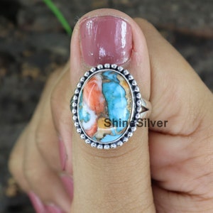 Peut inclure: Une bague en argent avec une pierre turquoise de forme ovale. La pierre pr&eacute;sente un m&eacute;lange de couleurs bleues, orange et blanches. La bague a une bordure perl&eacute;e.