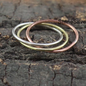 May include: Three thin rings in silver, gold, and rose gold are interlocked, set against a dark, textured background. The rings are simple bands, each with a smooth, polished finish. The rings are labeled with the text "ShineSilver".