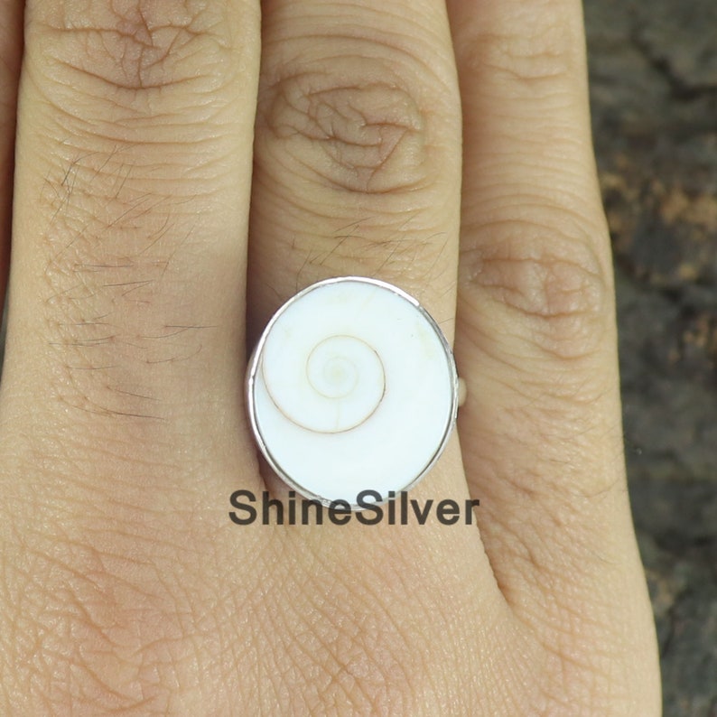 May include: A silver ring with a white shell inlay featuring a spiral pattern. The ring is on a finger.