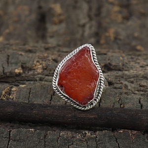 May include: A silver ring with a large, irregularly shaped, reddish-orange stone. The stone is set within a twisted rope-like border. The ring is displayed on a dark, textured wooden surface.
