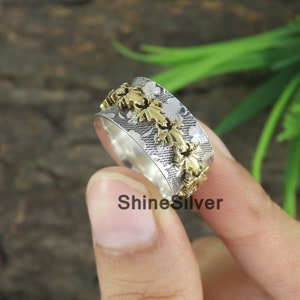 Könnte beinhalten: Ein Silber- und Goldring mit einem strukturierten Design und einer Reihe von goldenen Blattakzenten. Der Ring wird an einem Finger getragen.