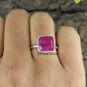 Könnte beinhalten: Ein silberner Ring mit einem quadratischen rosafarbenen Edelstein. Der Ring ist in einer Zargenfassung gefasst und hat eine polierte Oberfläche. Der Edelstein ist in einem leuchtenden Rosaton gehalten und hat einen leichten Glanz. Der Ring wird an einem Finger gezeigt.