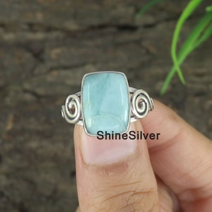 May include: A silver ring with a rectangular, light blue gemstone. The ring features a spiral design on each side of the gemstone. The text "ShineSilver" is visible on the ring.