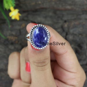 Peut inclure: Une bague en argent avec une pierre pr&eacute;cieuse ovale bleue sertie dans une b&eacute;li&egrave;re &agrave; perles. La bague est port&eacute;e sur un doigt.