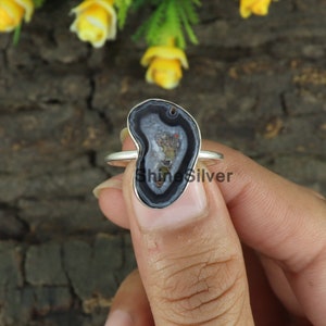 May include: A silver ring with an irregularly shaped agate stone. The stone displays a dark grey and white pattern with a hint of brown at the centre. The ring is held by a person's hand. The text "ShineSilver" is visible on the ring.