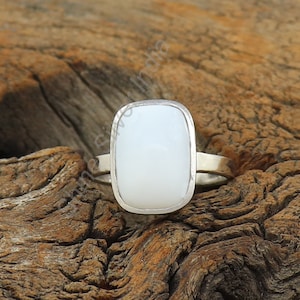 May include: A white oval gemstone ring set in a silver band. The ring is on a wooden surface.