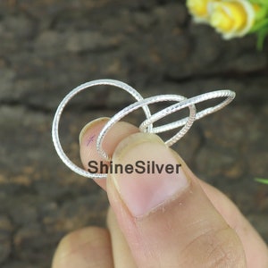 Könnte beinhalten: Ein silberner Ring mit einem einzigartigen Design, das drei miteinander verbundene, strukturierte Bänder aufweist. Der Ring hat eine helle, reflektierende Oberfläche. Der Text "ShineSilver" ist auf dem Ring zu sehen.