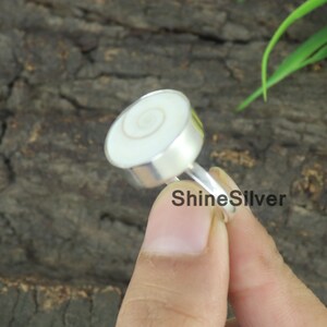 May include: A silver ring with a white shell inlay featuring a spiral pattern. The ring is on a finger.