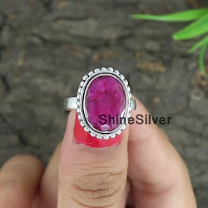 May include: A silver ring with a large, oval-shaped, pink ruby gemstone set in a beaded bezel. The ring is on a finger.