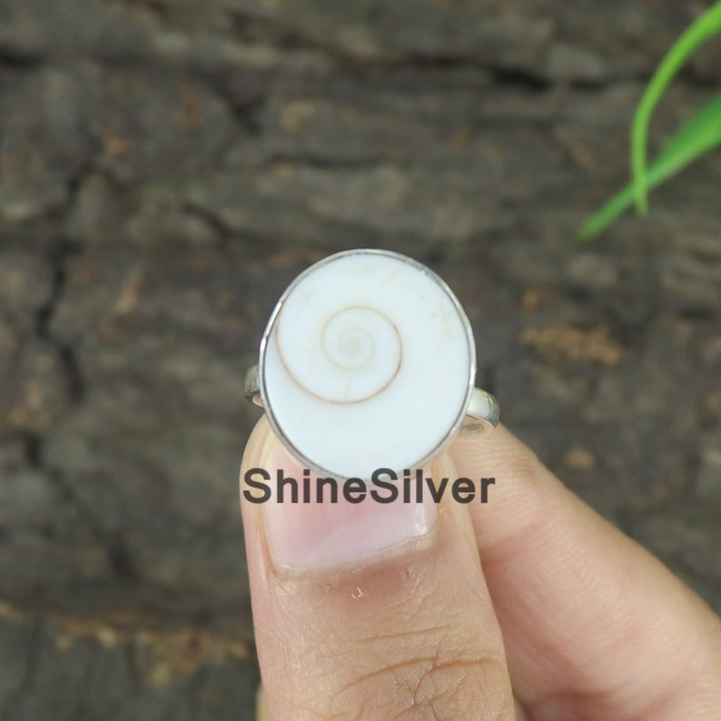 May include: A silver ring with a white shell inlay featuring a spiral pattern. The ring is on a finger.