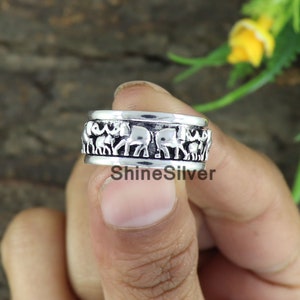 Könnte beinhalten: Ein silberner Ring mit einem drehbaren Band, das mit einem Muster von Elefanten verziert ist, die im Kreis laufen. Der Ring befindet sich an einem Finger.
