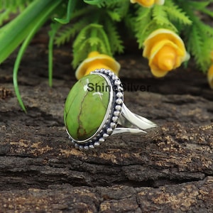 Peut inclure: Une bague en argent avec une pierre ovale verte sertie dans une b&eacute;li&egrave;re &agrave; perles. La bague est pos&eacute;e sur une surface en bois.