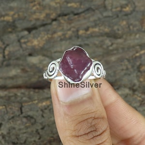 May include: A silver ring with a large, dark pink gemstone set in a swirl design. The ring is on a finger.