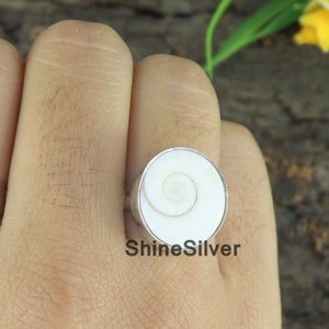 May include: A white shell ring with a silver band. The shell has a spiral pattern in the center.